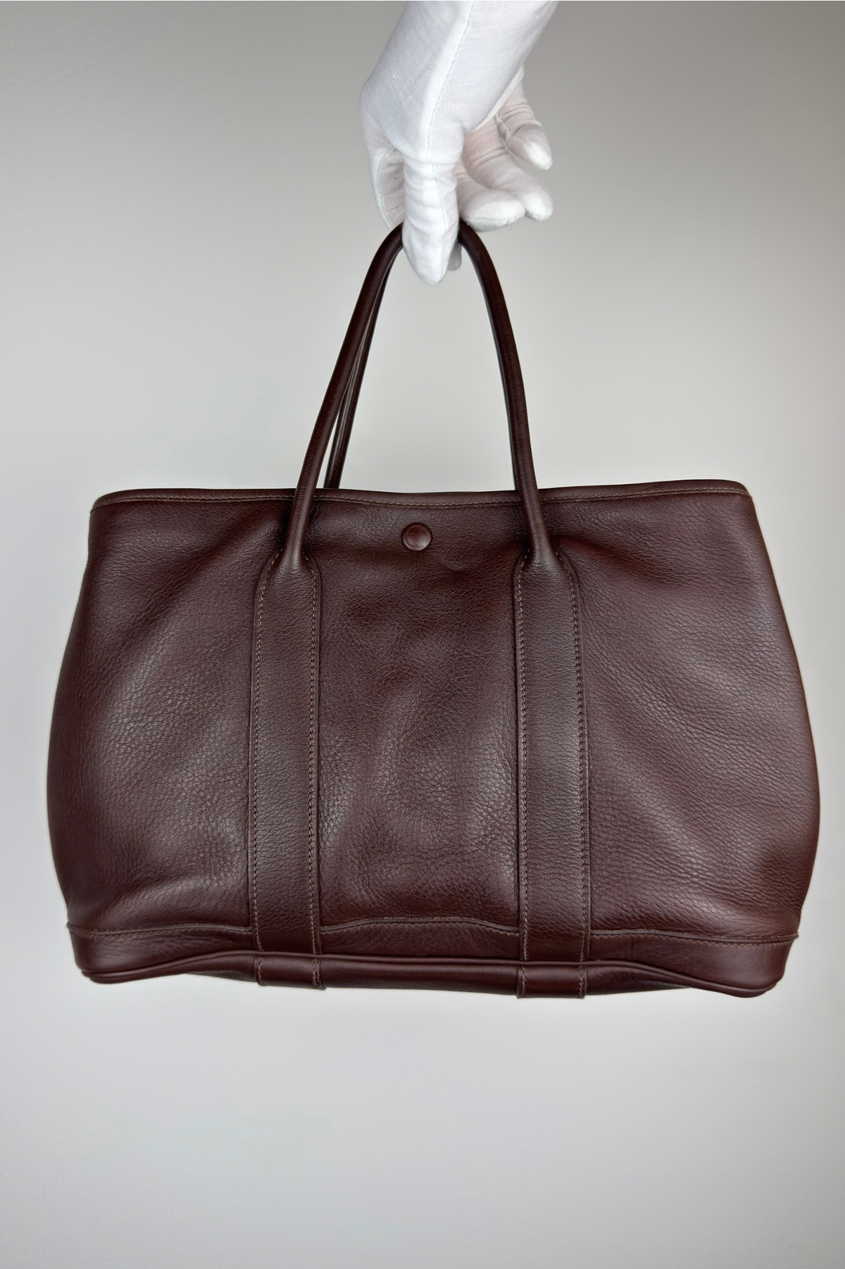 A dark brown leather shoulder bag with double top handles, held by a white gloved hand against a neutral light gray background. Hermès Garden Twilly 30 (TPM) in Veau Swift Leather (Color: Havana)